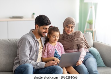 Young muslim parents and girl using laptop together at home, surfing internet or having video chat with someone, sittig on sofa. Happy arab man, child and woman in hijab staying home on isolation