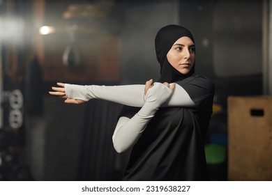 Young Muslim sportswoman in hijab and activewear stretching left arm while standing in front of camera during fitness workout in gym - Powered by Shutterstock