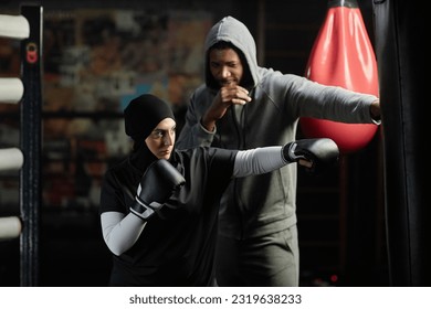 Young Muslim woman in activewear, hijab and boxing gloves hitting black leather punching bag while standing next to male instructor - Powered by Shutterstock