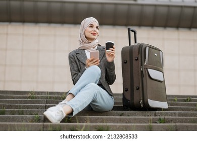 Young Muslim Woman In Hijab Dressed In Jeans And Jacket Using Phone And Drinking Take Away Coffee, While Sitting On The Stairs With Travel Bag.