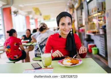 A Young Muslim Woman In A Hijab Head Scarf Eats Her Halal Nasi Lemak Lunch During The Day At A Table In A Hawker Center. 