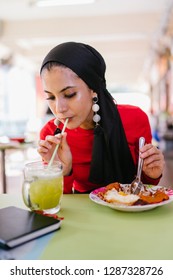 A Young Muslim Woman In A Hijab Head Scarf Eats Her Halal Nasi Lemak Lunch During The Day At A Table In A Hawker Center. 