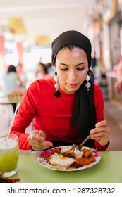 A Young Muslim Woman In A Hijab Head Scarf Eats Her Halal Nasi Lemak Lunch During The Day At A Table In A Hawker Center. 