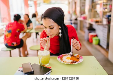 A Young Muslim Woman In A Hijab Head Scarf Eats Her Halal Nasi Lemak Lunch During The Day At A Table In A Hawker Center. 