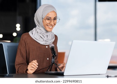 A young Muslim woman in a hijab is sitting in an office in front of a laptop and a microphone. records a podcast, a blog, talks on a video call, gives an interview, goes through an online interview. - Powered by Shutterstock