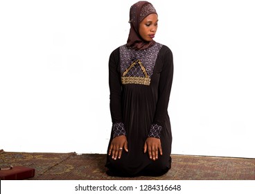 Young Muslim Woman Kneeling On A Mat Praying While Looking In Profile.