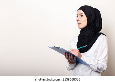 Young Muslim Woman In A White Shirt And Hijab Holds A Clipboard And Looks To The Side. Auditor Hiring. Banner.