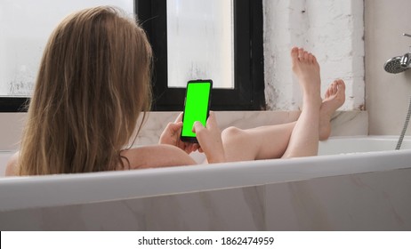 Young Naked Woman Lies In Light White Bathtub And Looks At The Phone With Green Screen. Lady Plays Swipe Up And Right On Cellular With Chroma Key With Legs Lay On Bath.