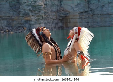 A Young Native American Indian Couple Stand Together In A Stone Quarry Lake. They Are Both Wearing Feathered Headdresses. The Young Indian Couple Are In Love With Each Other. 