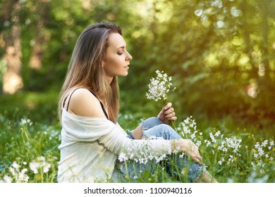 Young natural woman sitting in green grass among the trees in the sun with a bouquet of white flowers. Portrait in natural light and warm colors.
