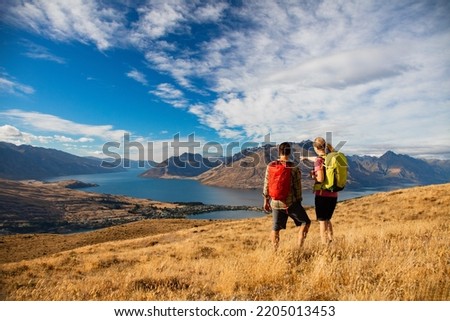 Young New Zealand travel tourist couple with backpacks relaxing viewing The Remarkables on vacation near Lake Wakatipu Otago