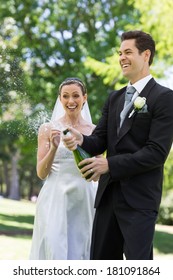 Young Newlywed Couple Popping Cork Of Champagne In Park