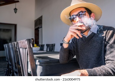 Young Nicaraguan man wearing a sweater hat and sunglasses smoking a cigar in the living room of a colonial-style mansion in Managua