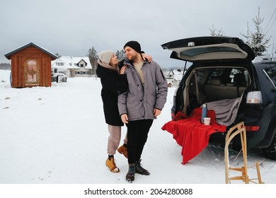 Young Nice Caucasian Couple Travel By Car In Winter. Cold Winter Clothing, Holidays,  Weekend At Countryside, Stay Outside, Drink Tea, Hug. Sitting On The Rear Part Of Modern Car, Have A Rest.