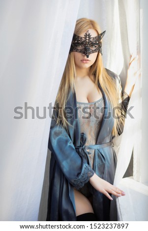 Young nice lady in a mask posing near the window, dressed in a bodysuit and bathrobe