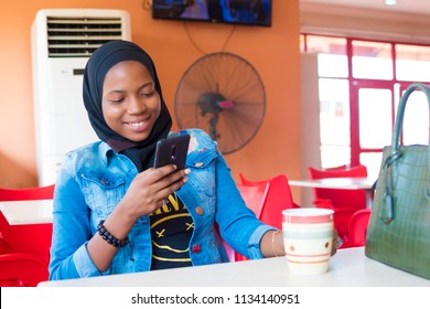 Young Nigerian Girl Smiling While Using Her Mobile Phone, Sitting Alone In A Fast Food Restaurant, With A Cup Of Tea Or Coffee 