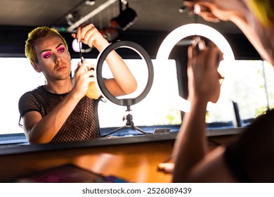 A young non binary with bright makeup adjusts a ring light. The non binary with vibrant makeup is preparing for a shoot. The non binary is focused on the ring light setup while doing makeup. - Powered by Shutterstock