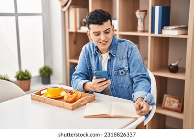 Young non binary man using smartphone and writing on notebook having breakfast at home