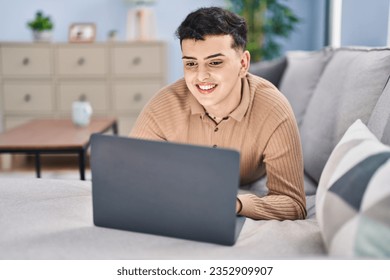 Young non binary man using laptop lying on sofa at home