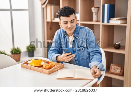 Young non binary man using smartphone and writing on notebook having breakfast at home