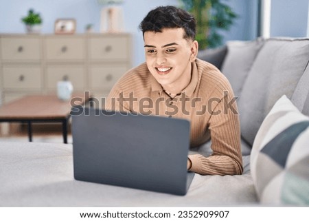 Young non binary man using laptop lying on sofa at home