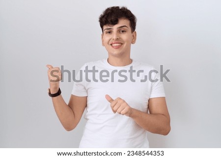 Young non binary man wearing casual white t shirt pointing to the back behind with hand and thumbs up, smiling confident 