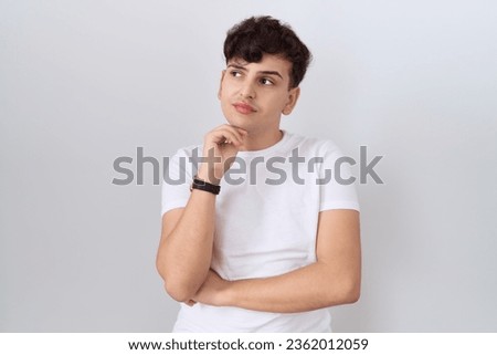 Young non binary man wearing casual white t shirt with hand on chin thinking about question, pensive expression. smiling with thoughtful face. doubt concept. 