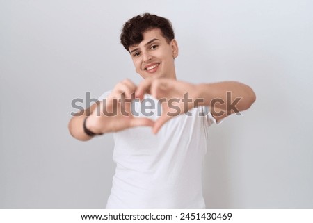 Young non binary man wearing casual white t shirt smiling in love doing heart symbol shape with hands. romantic concept. 
