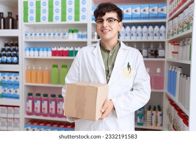 Young non binary man working at pharmacy drugstore holding box looking positive and happy standing and smiling with a confident smile showing teeth 