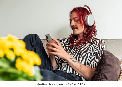 Young non-binary adult with pink hair and tattoos wearing zebra shirt and headphones is lying on sofa using smartphone, listening to music and enjoying free time at home