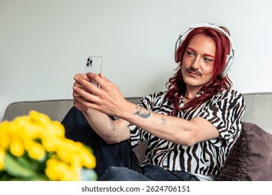 Young non-binary influencer with red hair and headphones taking a selfie with smartphone sitting on the sofa at home, with yellow flowers in the foreground
