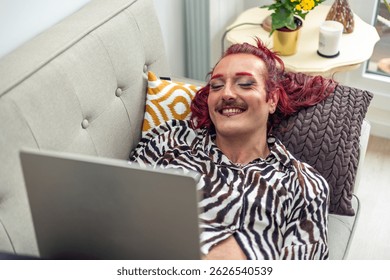 Young non-binary person with red hair and makeup smiling and using laptop on comfortable sofa at home, enjoying modern technology and celebrating body positivity and gender identity