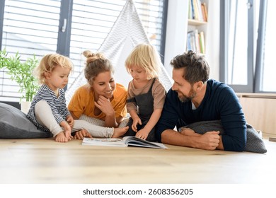 Familia nuclear joven jugando con juguetes en una sala de estar. Padres y niños acostados en el suelo, mirando el libro de cuentos de los niños, pasando el día de fin de semana en interiores.