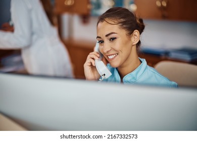 Young Nurse Answering A Phone Call While Working On Desktop PC At Reception Desk In A Hospital. Copy Space.