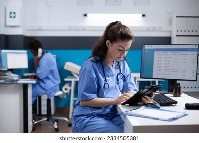 Young nurse checking appointments list on digital tablet in busy medical office. Adult woman healthcare specialist working with technology at hospital desk with stethoscope around neck - Powered by Shutterstock