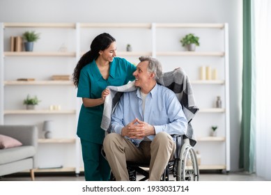 Young nurse covering disabled elderly man in wheelchair with warm plaid at retirement home. Millennial caregiver assisting handicapped senior patient, taking care of older male indoors