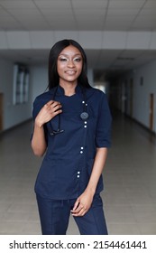 Young Nurse In Dark Blue Uniform And Stethoscope Over Her Neck. Portrait Of Female Nurse Wearing Scrubs In Hospital. Female Nurse Or Doctor Smiles Looking To Camera In Hospital Hallway