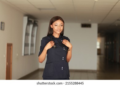 Young Nurse In Dark Blue Uniform And Stethoscope Over Her Neck. Portrait Of Female Nurse Wearing Scrubs In Hospital. Female Nurse Or Doctor Smiles Looking To Camera In Hospital Hallway