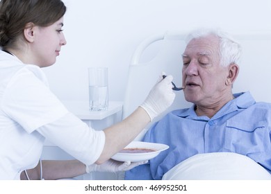 Young Nurse Feeding The Senior Patient Lying On A Hospital Bed