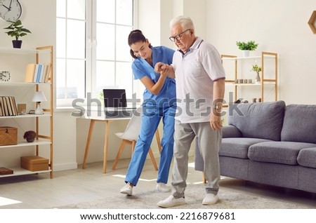 Young nurse helping elderly man walk in the room, holding his hand, supporting him. Treatment and rehabilitation after injury or stroke, life in assisted living facility, senior care concept