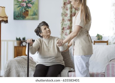 Young Nurse Helping Sad Elderly Woman To Stand Up From The Sofa