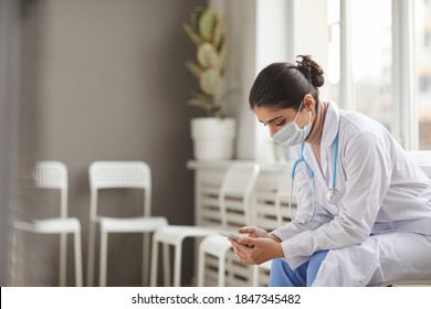 Young Nurse In Mask And In White Coat Using Her Mobile Phone During Her Break Sitting On Chair At The Corridor