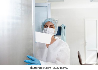 A Young Nurse In A Protective Suit, Cap, Mask And Gloves Takes Documents From A Locker In The Medical Office