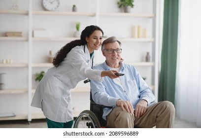 Young Nurse With Remote Control Switching Channels On TV For Senior Man In Wheelchair At Retirement Home. Millennial Caregiver Assisting Mature Male Patient With Disability Indoors