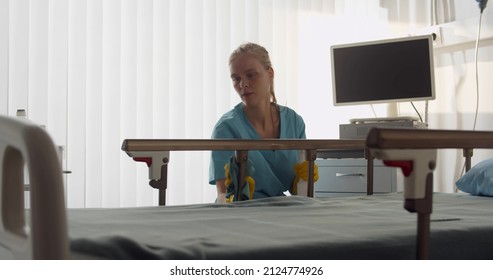 Young Nurse In Scrubs Cleaning Bed With Detergent Sprayer In Hospital Ward. Female Medical Worker Disinfecting Furniture In Clinic Room. Healthcare And Hygiene Concept