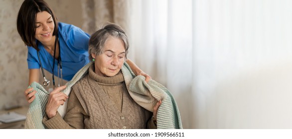 A young nurse takes care of an elderly 80-year-old woman at home, wraps a blanket around her. Happy retired woman and trust between doctor and patient. Medicine and healthcare.