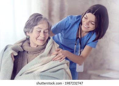 A Young Nurse Takes Care Of An Elderly 80-year-old Woman At Home, Wraps A Blanket Around Her. Happy Retired Woman And Trust Between Doctor And Patient. Medicine And Healthcare.