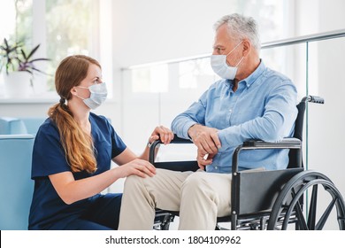 Young Nurse Taking Care Of Mature Male Patient On Wheelchair In Hospital. Caucasian Woman And Old Man Wearing Face Mask For Protection Of Covid 19 Pandemic