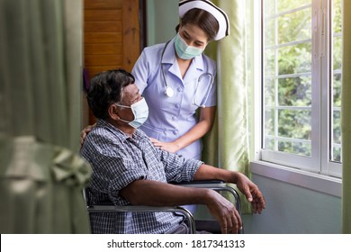 Young Nurse Wearing Medical Protective Mask Taking Care Of An Elderly Patient Sitting In A Wheelchair In Hospital Room.