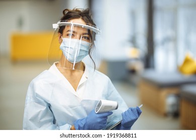 Young Nurse Wearing Medical Protective Face Shield And Mask Stands At The Hospital Corridor With A Non Contact Thermometer And Tablet Ready To Test The Patients. Health Care And Diagnosis Concept